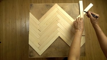 Measuring and cutting paint stir sticks to tabletop to create herringbone pattern.