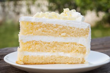 Vanilla sponge cake with cream and white chocolate decorate. Sliced piece of cake on white plate. Served on wooden table.