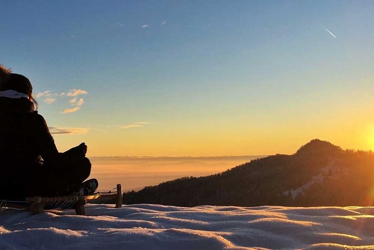 Young woman sitting and meditating on sledge looking at sunrise in winter.