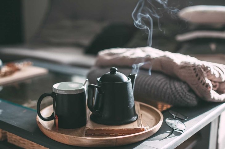 Still life details in home interior of living room. Sweaters and cup of tea with steam on a serving tray on a coffee table. Breakfast over sofa in morning sunlight. Cozy autumn or winter concept.