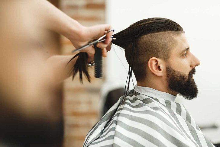 Man's hands doing a haircut for man with dark long hair and beard at barber shop, close up portrait, copy space.